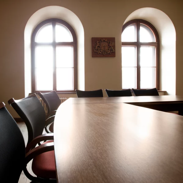 Tukša sanāksmju telpa ar garu koka galdu un krēsliem. Divi gaiši arkveida logi apgaismo telpu, un starp tiem pie sienas ir koka emblēma.Kuldīgas novada pašvaldības mazā zāle.