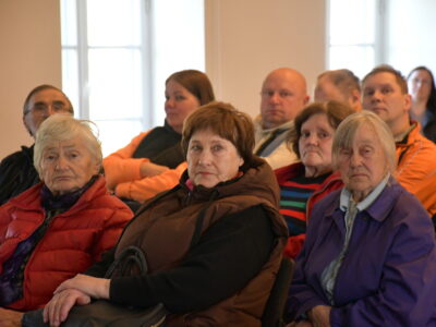 Cilvēku grupa, galvenokārt gados vecāki cilvēki, sēž iekštelpās, klausoties vai vērojot priekšā notiekošo. Priekšplānā redzamas trīs gados vecākas sievietes: viena ar baltiem matiem sarkanā jakā, otra ar brūniem matiem brūnā vestē, trešā ar sirmiem matiem violetā jakā.
