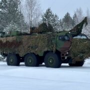Bruņots zaļš militārais transportlīdzeklis ar maskēšanās tīklu stāv uz sniega klātas zemes ziemas mežā.