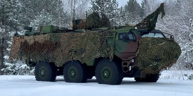 Bruņots zaļš militārais transportlīdzeklis ar maskēšanās tīklu stāv uz sniega klātas zemes ziemas mežā.