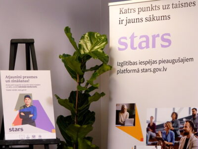Divi plakāti reklamē izglītības platformu pieaugušajiem "stars.gov.lv". Kreisais plakāts ar uzrakstu "Atjaunini prasmes un zināšanas!" attēlo smaidīgu vīrieti un tekstu "Virzies pretī izaugsmei vai jaunām darba iespējām! Lai sāktu, izveido individuālo mācību kontu. Uzzini vairāk: stars.gov.lv". Labais plakāts, kurā lieli burti veido vārdu "stars", satur uzrakstus "Katrs punkts uz taisnes ir jauns sākums" un "Izglītības iespējas pieaugušajiem platformā stars.gov.lv", kā arī attēlus ar cilvēkiem, kas mācās vai strādā ar datoru. Starp plakātiem stāv liels zaļš telpaugs.