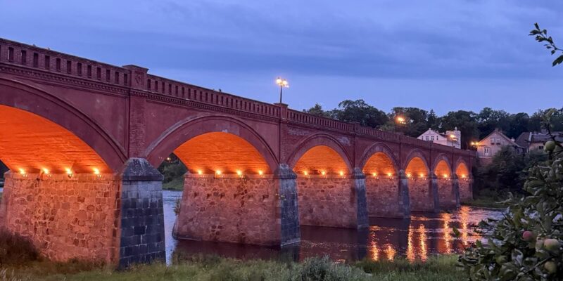 Vecs sarkano ķieģeļu velvju tilts ar akmens balstiem, kas krēslā izgaismots ar silti oranžām gaismām, atspoguļojas upē. Priekšplānā zāļaini krasti.