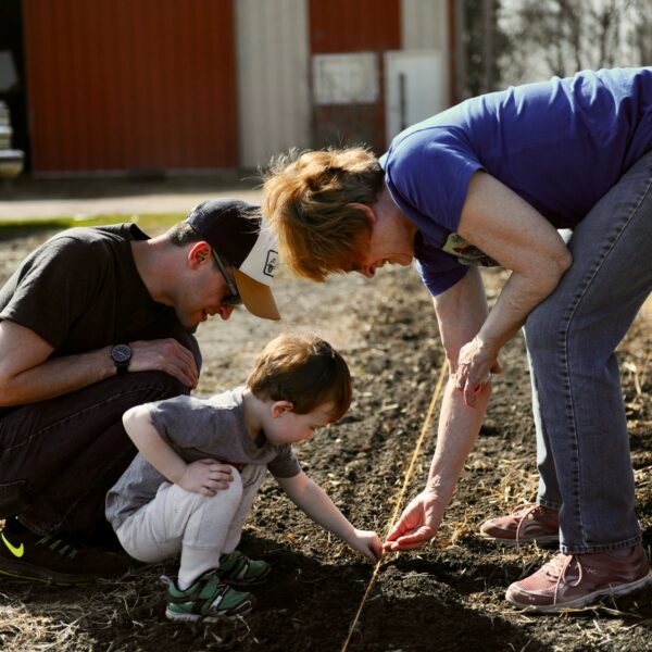Trīs cilvēki – vīrietis, mazs bērns un sieviete – tupus stāda sēklas augsnē, izmantojot auklu kā vadlīniju. Sieviete rāda bērnam, kā to darīt. Saules apgaismotā fonā redzams sarkans šķūnis un kravas automašīna.